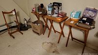Four light oak folding tables aligned against a wall, standing on a basement floor with visible stains. Various objects on table tops not included.