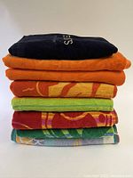 Stack of six folded beach towels in various colors and patterns including navy, solid orange, patterned orange, green, red, and multicolor towels.