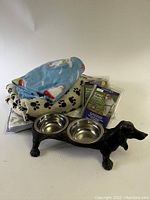 Photo showing the dachshund cast iron dog bowl with two stainless steel bowls in place, two packaged Easy Walk small dog leashes, and several dog bedding blankets stacked behind.