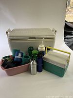 Photo showing a large off-white Igloo Marine cooler with side handles, a smaller teal and white Coleman cooler with yellow handle, multiple assorted water bottles and colorful bottle sleeves in a pink tub.