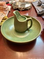 Green ceramic pitcher inside green ceramic bowl on wooden surface with textiles in background