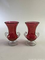 Front view of two cranberry colored glass vases with clear glass handles and round pedestal bases on white background