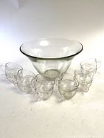 Large transparent glass punch bowl with 12 matching cups arranged around it on white surface.