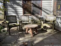 Four wooden patio chairs and one small round wooden table on patio. Chairs are vertical slat style with armrests, table is low and round with three legs.