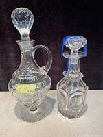 Two vintage glass decanters shown side by side on a light surface against a dark background. The decanter on the left is taller with a handle and a round faceted stopper. The decanter on the right is shorter, hexagonal with a faceted geometric stopper.