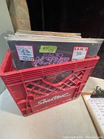 Photo of red plastic crate labeled 'Sealtest' containing multiple vinyl records, with some album covers visible including one marked Radio Active.