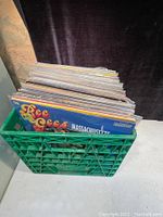 Green plastic milk crate filled with a collection of vinyl records, showing edges of album covers including The Bee Gees Massachusetts.