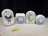 Photo showing four plug-in mechanical security timers with dials and manual switches placed on a neutral surface.