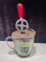 Front view of antique hand-crank beater with original ribbed glass pitcher, metal crank with large gear and red wooden handle.