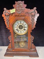 Full front view of antique oak gingerbread clock showing ornate carved floral and leaf details on case and clock face with pendulum visible behind glass.