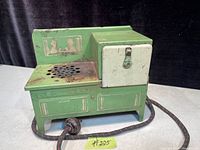 Green and cream Art Deco electric kids toy stove with vented stovetop and small oven door, showing signs of age wear and rust on metal surface. Original fabric-covered power cord visible.