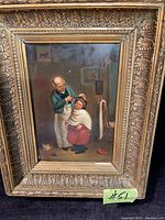 Front view of the oil painting showing the scene of an older man cutting a child's hair with a cat nearby, inside a gilded wood frame.