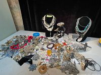 Wide view of entire lot showing many silver and gold-tone costume jewelry pieces including necklaces, chains, brooches, earrings, and watches on a white surface with black display busts.