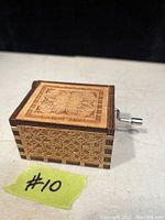 Closed view of the small wooden rectangular musical box showing detailed carved patterns on all sides and the lid. Metal hand crank on the side is visible.