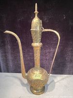 Full front view of the tall vintage solid brass Persian coffee pot showing the elongated spout, handle, and pointed finial lid with engraved floral patterns.