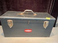 Closed vintage blue metal toolbox with Tecomaster label, showing metal clasps and handle with some rust and wear.
