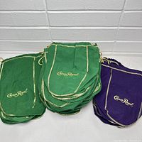 Photo shows stacks of green and purple Crown Royal whiskey bottle bags laid on a grey surface against white wall.