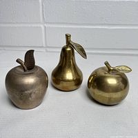 Three decorative fruit figurines showing brass pear, brass apple, and silver-toned apple against a white background.