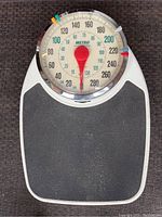 Top view showing large circular dial with red needle pointer, five colored markers, black textured standing platform on white base.