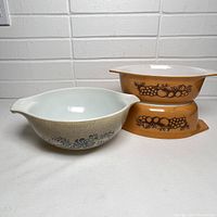 Two Pyrex casseroles stacked, showing fruit motif design on mustard yellow background and a third bowl with white interior and floral pattern.