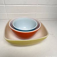 Three vintage Pyrex bowls nested together showing the color and size difference: yellow square bowl, large orange round bowl, smaller blue round bowl inside.