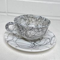 Tea cup seated on saucer showing outside and inside black spiderweb spaghetti pattern on white milk glass.