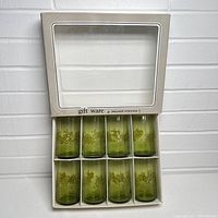 Box with eight green glasses visible through clear window, showing the original packaging with some wear.