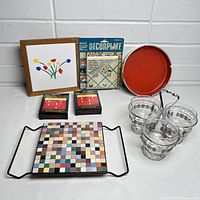 Photo of the full lot showing all included items arranged on a white surface against a white wall, including mosaic tile tray, glass set with holder, framed floral art, boxed candles, red tray, and craft kit