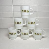 Front and side view of 6 white milk glass Fire King mugs arranged in a pyramid with visible green and yellow floral decoration around the exterior.