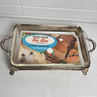 Top down view showing the rectangular glass baking dish with intact Fire King label inside the metal stand with decorative handles and claw feet.