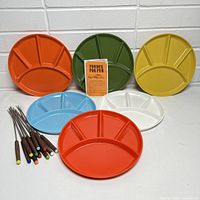 Full overview of the 6 solid color 9-inch vintage fondue plates arranged in two rows, with 11 teak handle fondue forks with colored tips laid out in front and a Fondue For Fun pamphlet in the middle.
