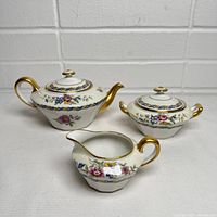 Front view of the complete tea set showing teapot, creamer, and sugar bowl with floral design and gold accents