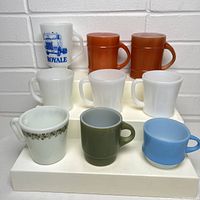 Front-facing photo showing all nine vintage milk glass mugs arranged on a three-tiered white display with a white brick background