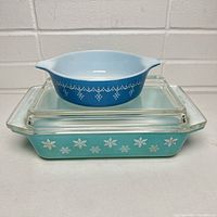 Photo shows stacked large rectangular turquoise blue Pyrex baking dish with white snowflake pattern and clear glass lid, topped with small blue round Pyrex dish with white pattern.
