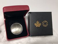 Silver coin in red protective case next to black Royal Canadian Mint box with gold logo