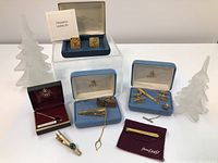 Overview of multiple men's accessories including cufflinks and tie clips in various boxes and display pouches, staged with decorative frosted Christmas tree figures in background.