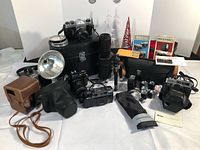 Overall lot view showing Pentax SLR camera on case, Minolta Hi-Matic F camera, lenses, flash units, tripod, Ricoh camera, MagicCube bulbs