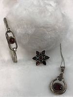 Photo showing garnet bracelet, flower-shaped ring with garnets, and chain with garnet pendant on white plush background.