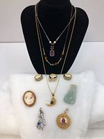 Full lot of jewelry showing three necklaces displayed on black stand along with three rings and various pendants and brooches on white surface below