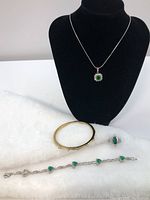 Jewelry lot displayed on black stand and white surface including necklace with green stone pendant, gold-tone bangle, silver ring, and silver bracelet with green heart stones.