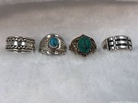 Top view of four sterling silver rings arranged on white cloth background: chain inlay band, turquoise bezel ring, carved turquoise ornate ring, geometric pattern ring.