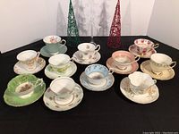 Front view of nine vintage tea cup and saucer sets arranged on a table with holiday decoration cones behind.