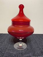 Full view of the ruby red Empoli glass jar with circus tent lid, showing the smooth glossy finish and clear glass pedestal base