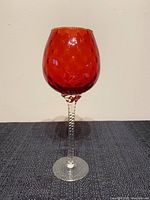 Full view of the mid century ruby red glass balloon snifter with twisted clear glass stem standing upright on a dark surface against a neutral background showing its height and form.