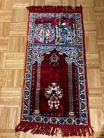 Full view of the red prayer rug carpet with architectural motifs and fringed edges laid flat on a wooden floor.