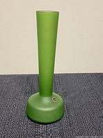 Front view of tall green frosted glass vase standing upright on a textured dark surface, showing shape and color clearly.