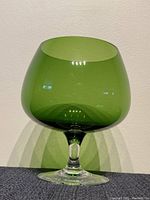 Front view of vintage green glass brandy snifter with clear stem and foot on a dark surface against a light wall.