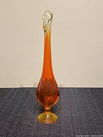 Front view of a tall orange and yellow Mid Century Modern glass vase with ruffled clear glass top and bulbous base on a dark textured surface.