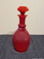 Red pressed glass decanter bottle with diamond texture, shown on black textured cloth against beige wall.