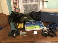Image showing camping tent, stakes, tripod grill box, cooler, flashlight, water pump device, and bags on wooden table.